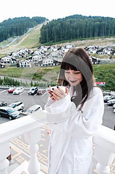 Beautiful shy girl in white hotel bathrobe drinking hot coffee on a balcony. Nature and nice view outside. Quiet weekend getaway
