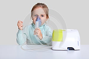 Beautiful sick girl has inhalation therapy, uses portable nebulizer, holds mask vapor, sits at white desktop, isolated over studio