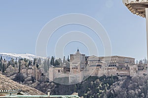 Beautiful side view of the Alhambra on a hill with arid terrain and some green trees with a snowy mountain in the background