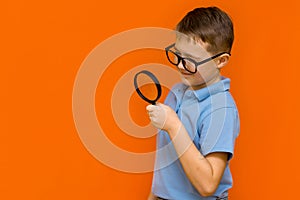 Beautiful side view european young boy with glasses holding and looking through magnifying glass.Orange wall