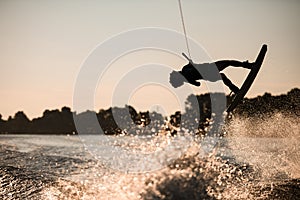 Beautiful silhouette of female rider holding rope and making jump on wakeboard at sunset. Water sports activity.