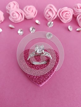 Beautiful silver and diamond engagement ring on a pink heart with pink roses and loose diamonds on a pink background