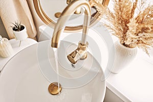 A beautiful sink with water turned on with a golden faucet next to an oval mirror and a shelf with hand towels. Close-up