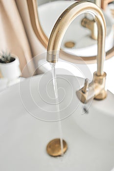 A beautiful sink with water turned on with a golden faucet next to an oval mirror and a shelf with hand towels. Close-up