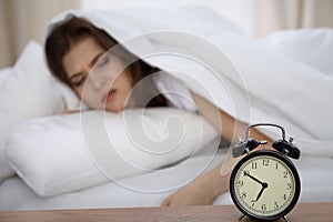 Beautiful sleeping woman lying in bed and trying to wake up with alarm clock. Girl having trouble with getting up early