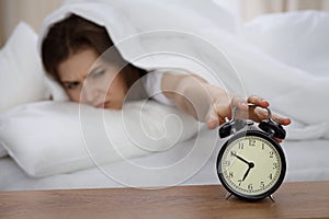 Beautiful sleeping woman lying in bed and trying to wake up with alarm clock. Girl having trouble with getting up early