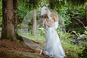Beautiful slender blond girl in dress hugging a gray horse, outdoors in summer in the forest