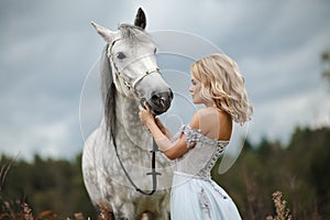 Beautiful slender blonde girl in dress looks at gray horse, on n