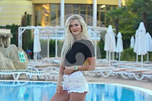 A beautiful slender fitness athletic blonde girl in white shorts and a black short T-shirt, standing by the pool at the hotel in