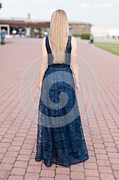 Slender blonde girl in dark-blue floor-length dress turned her back on the pavement
