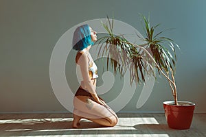 Beautiful slender girl in panties and a T-shirt with blue hair posing on the floor next to a flower in a pot
