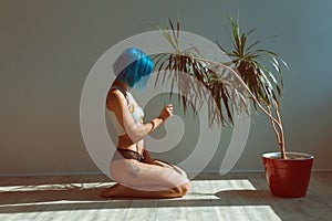 Beautiful slender girl in panties and a T-shirt with blue hair posing on the floor next to a flower in a pot