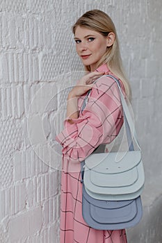 A beautiful slender girl in a pink dress poses against a white wall, she holds two handbags in her hands