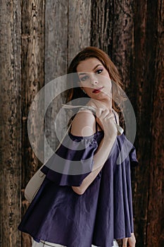 A beautiful slender girl in a short summer purple blouse, with chic makeup and flowing hair stands against a wooden wall