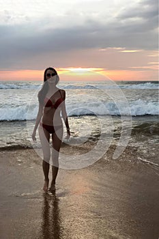 Beautiful slender tall young woman walking in a swimsuit against the ocean with the sunset.