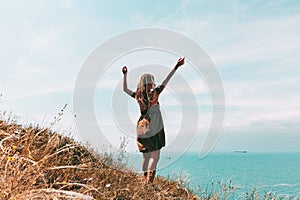 Beautiful slender tattooed woman with blonde dreadlocks in a dress rejoices on a hill. Sea View Travel and Vacation lifestyle