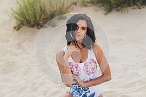 A beautiful slender young European woman sits on white sand, laughs and smiles. The girl is dressed in a fashionable