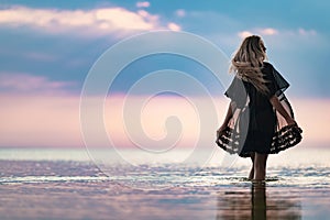 A girl with blond chic hair in a black bikini and a lace shawl walks along the estuary