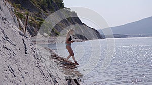 beautiful slender young woman in blue and black swimsuit enters sea or ocean on shore with rocks. pretty blonde girl
