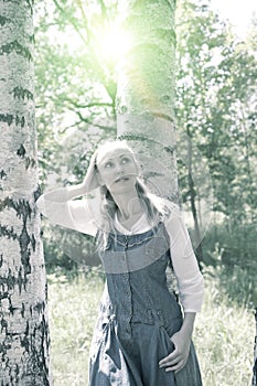 Beautiful slender young woman in a blue sundress and a white blouse near a birch in sunny day, toning
