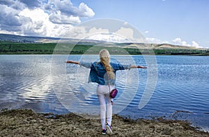 A beautiful slender young woman stands with her back on the shore near the water and admires the beautiful landscape