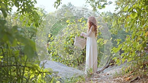 Beautiful slim caucasian woman with red hair standing on the riverbank holding basket and touching her long hair