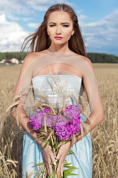 Beautiful slim girl in a blue dress in the field with a bouquet of flowers and ears of corn in his hands at sunset on a sunny