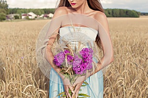 Beautiful slim girl in a blue dress in the field with a bouquet of flowers and ears of corn in his hands at sunset on a sunny