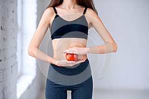Beautiful slim girl in dark sportwear holding red apple in the hands near her attractive belly. Healthy lifestyle, diet