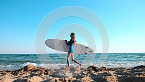 Beautiful slim girl walking on the beach, holding surfboard. Runs in the evening during sunset. Wears a swimming suit