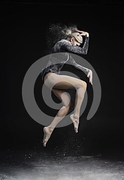 Beautiful slim girl wearing a black gymnastic bodysuit covered with clouds of the flying white powder jumps dancing on a dark.