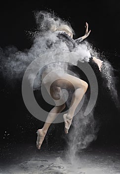 Beautiful slim girl wearing a black gymnastic bodysuit covered with clouds of the flying white powder jumps dancing on a dark.