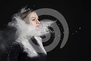 Beautiful slim girl wearing a black gymnastic bodysuit covered with clouds of the flying white powder poses on a dark background.