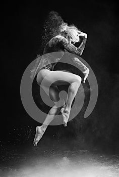 Beautiful slim girl wearing a gymnastic bodysuit covered with clouds of the flying white powder jumps dancing on a dark. Artistic