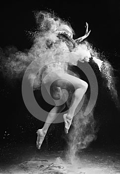Beautiful slim girl wearing a gymnastic bodysuit covered with clouds of the flying white powder jumps dancing on a dark. Artistic