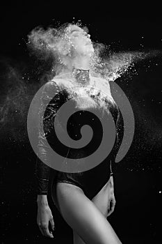 Beautiful slim girl wearing a gymnastic bodysuit covered with clouds of the flying white powder poses on a dark background.