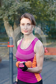 Beautiful slim and sporty girl in bright sportswear trains with dumbbell for biceps at outdoor sportground. White earphones, prote