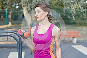 Beautiful slim and sporty girl in bright sportswear trains with dumbbell for biceps at outdoor sportground. White earphones, prote