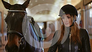 Beautiful slim woman in helmet and leather jacket walking in stable pulling horse bridle. Front view dolly shot portrait