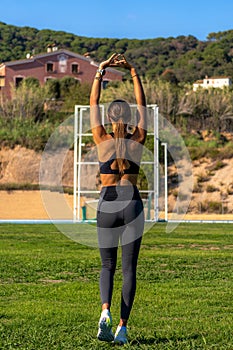 Beautiful slim young runner girl, tanned with long dark hair, stretching her back with her arms stretched above her head before