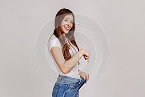 Beautiful slim young woman showing successful weight loss, pointing at big jeans.