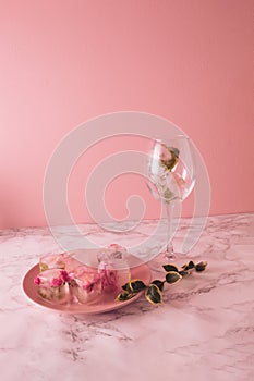 Beautiful small pink flowers in ice cubes and glass with petals on pink plate on marble and pink background