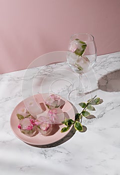 Beautiful small pink flowers in ice cubes and glass with petals on pink plate on pink background