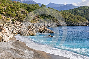 Beautiful small rocky beach with clear blue sea, waves and mountains on background. Oludeniz, Turkey