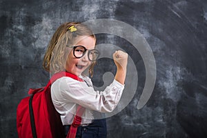 Beautiful smart little girl showing power muscles. Back to school. Kid in glasses with bag