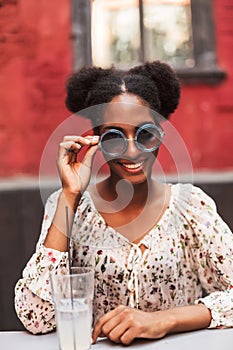 Beautiful smiling african lady in blouse and sunglasses while happily looking in camera with cocktail in cozy courtyard