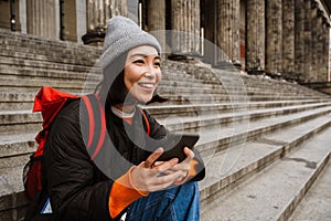 Beautiful smiling asian woman using smartphone while sitting on stairs at old city street