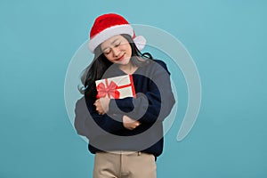 Beautiful smiling Asian woman wearing warm sweater and Christmas hat hugging red gift box  on blue background