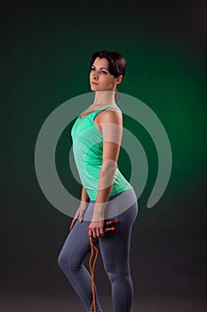 Beautiful smiling athletic, fitness woman standing, posing with a jump rope on a gray background with a green backlight