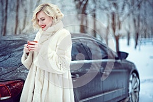 Beautiful blond woman in luxurious white fur coat drinking hot coffee on snowy winter day photo